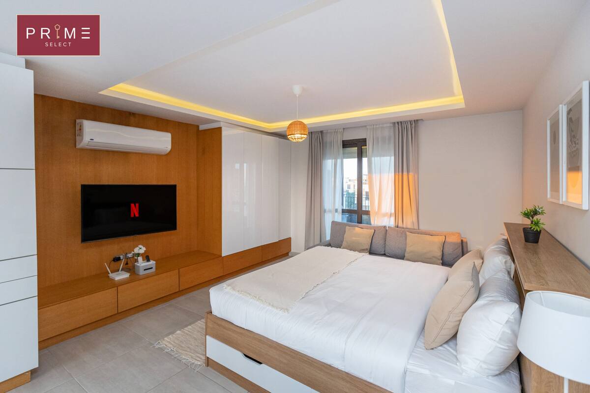 A modern bedroom with a large bed, a TV mounted on the wall, a wooden cabinet, and a bedside table with a plant.