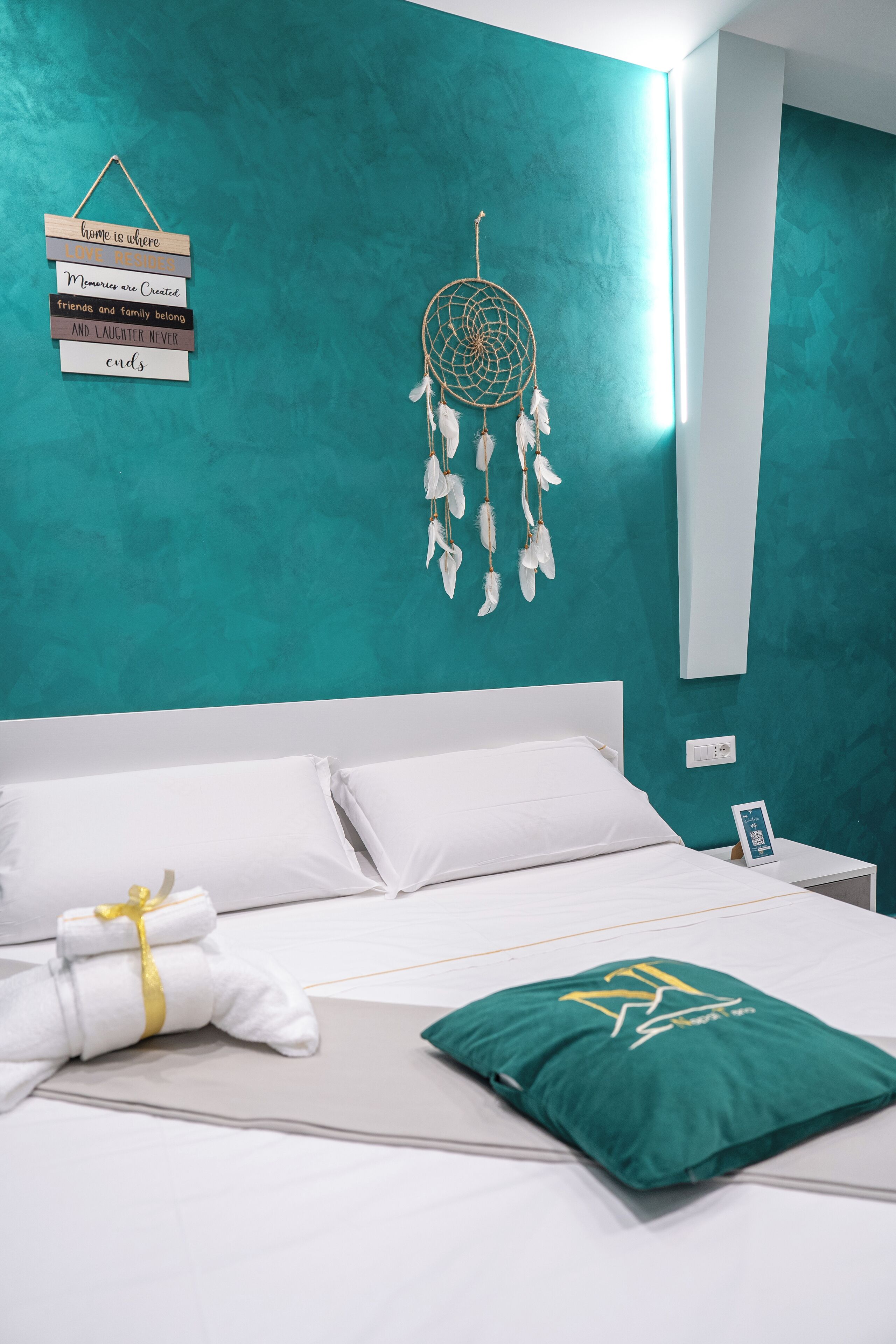 Een net opgemaakt bed met witte lakens en een groen kussen, een dreamcatcher aan de muur, en een bord met inspirerende citaten.