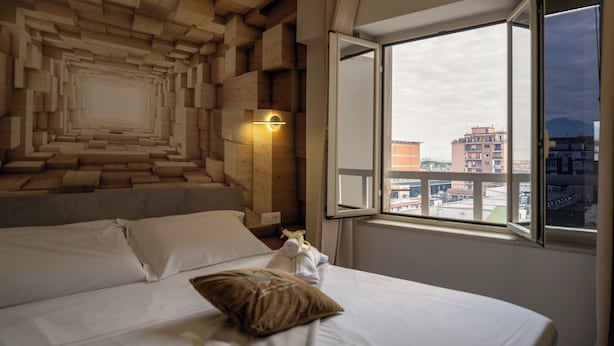 Une chambre d’hôtel dotée d’un décor mural en bois original, d’une grande fenêtre offrant une vue sur la ville et d’un lit soigneusement fait avec des oreillers.