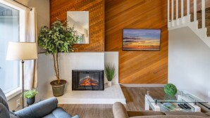 A modern living room with a fireplace, wooden wall paneling, a staircase, and a large window.