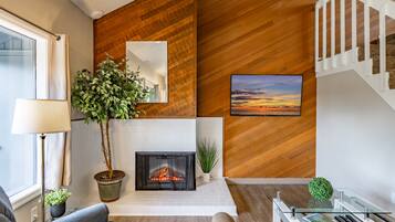 A modern living room with a fireplace, wooden wall paneling, a staircase, and a large window.