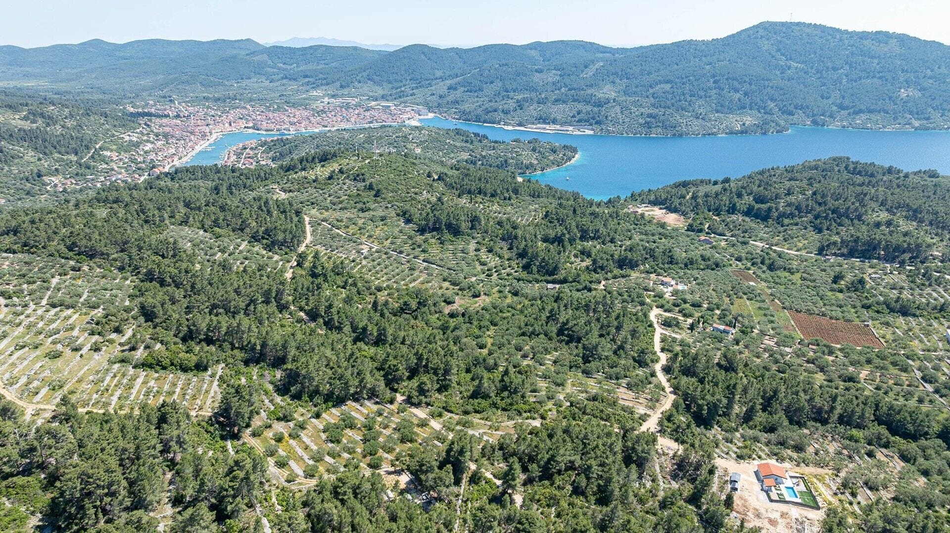 Ovenudblik over en kystby med et tæt skovområde, en klar blå sø og et lille boligområde med swimmingpool.