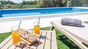 A poolside setting with a table, two glasses of orange juice, a bowl of nuts, and a towel on a lounge chair.