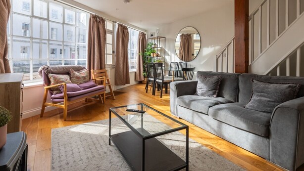 A modern living room with a grey sofa, a glass coffee table, and a wooden floor. There is a dining table with chairs and a staircase in the background. - Bright & Quirky 3BD Home - Wellington Terrace! (Bristol)