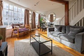 Een moderne woonkamer met een grijze bank, een glazen salontafel en een houten vloer. Er staat een eettafel met stoelen in en een trap op de achtergrond.