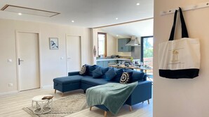 A modern living room with a blue sectional sofa, a small round table, and a kitchen in the background. - Le Châlet, Confort à L'orée de la Forêt et la mer (Mimizan)