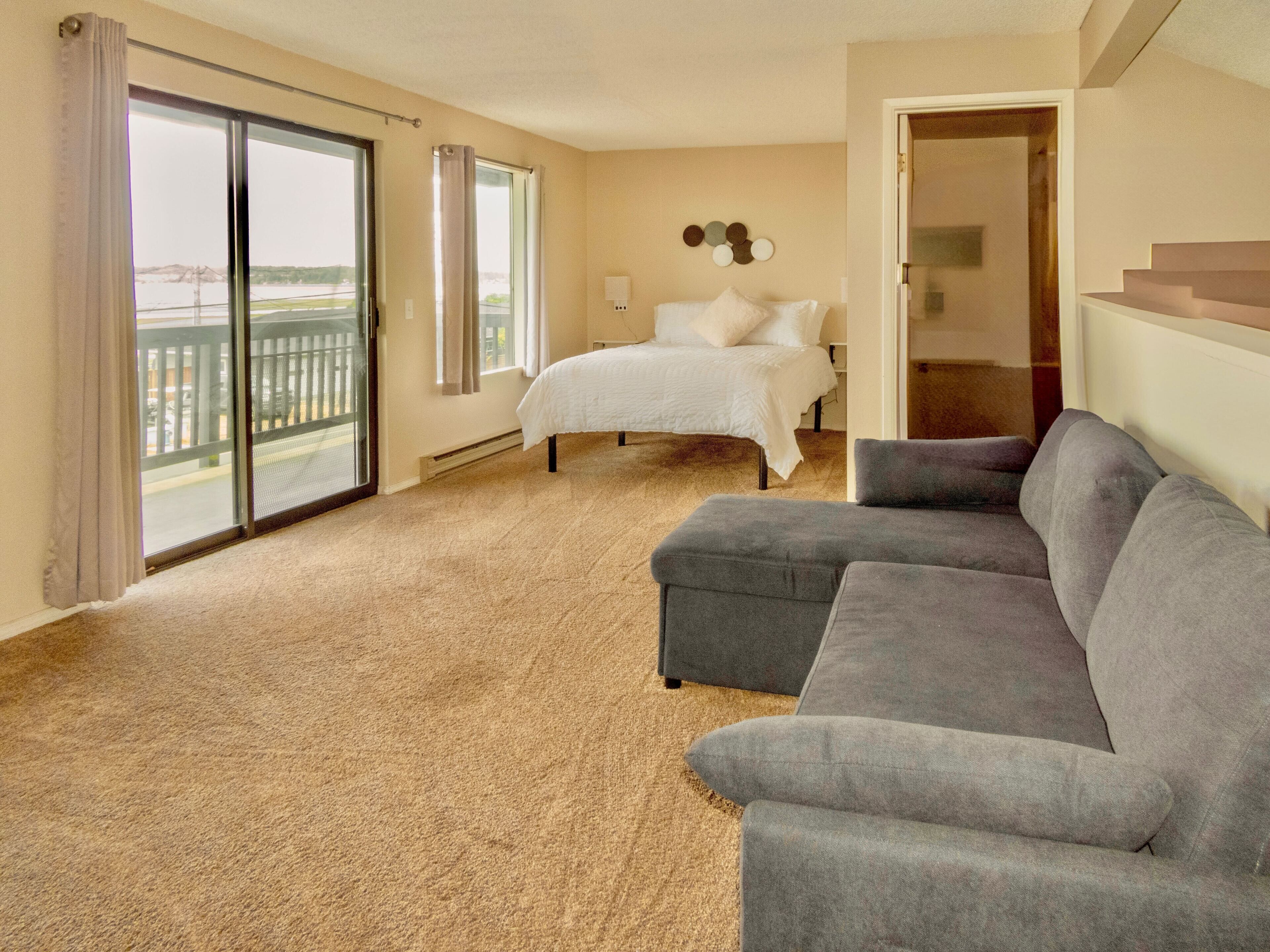 A hotel room with a large bed, a gray sofa, a balcony with a view, and a door leading to another room.