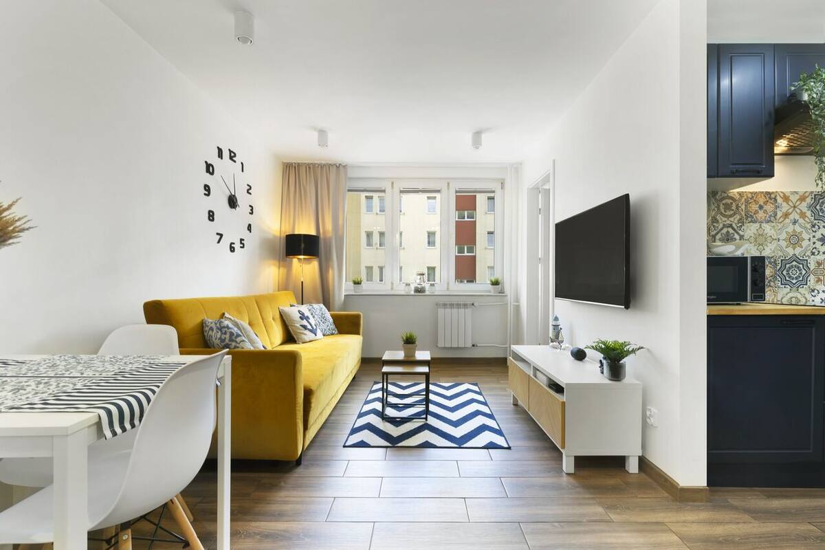 A modern living room with a sofa, a dining table, and a TV. There is a kitchen area with cabinets and a window.