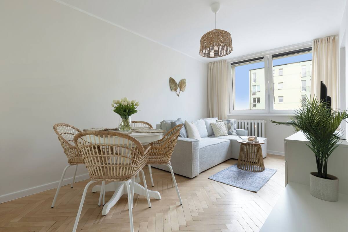 A modern living room with a sofa, dining table, wicker chairs, a hanging light fixture, and a potted plant.