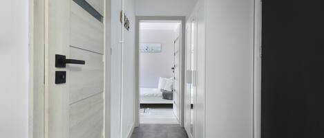 A narrow hallway with a bed in the background, a door on the right, and a light switch on the wall.