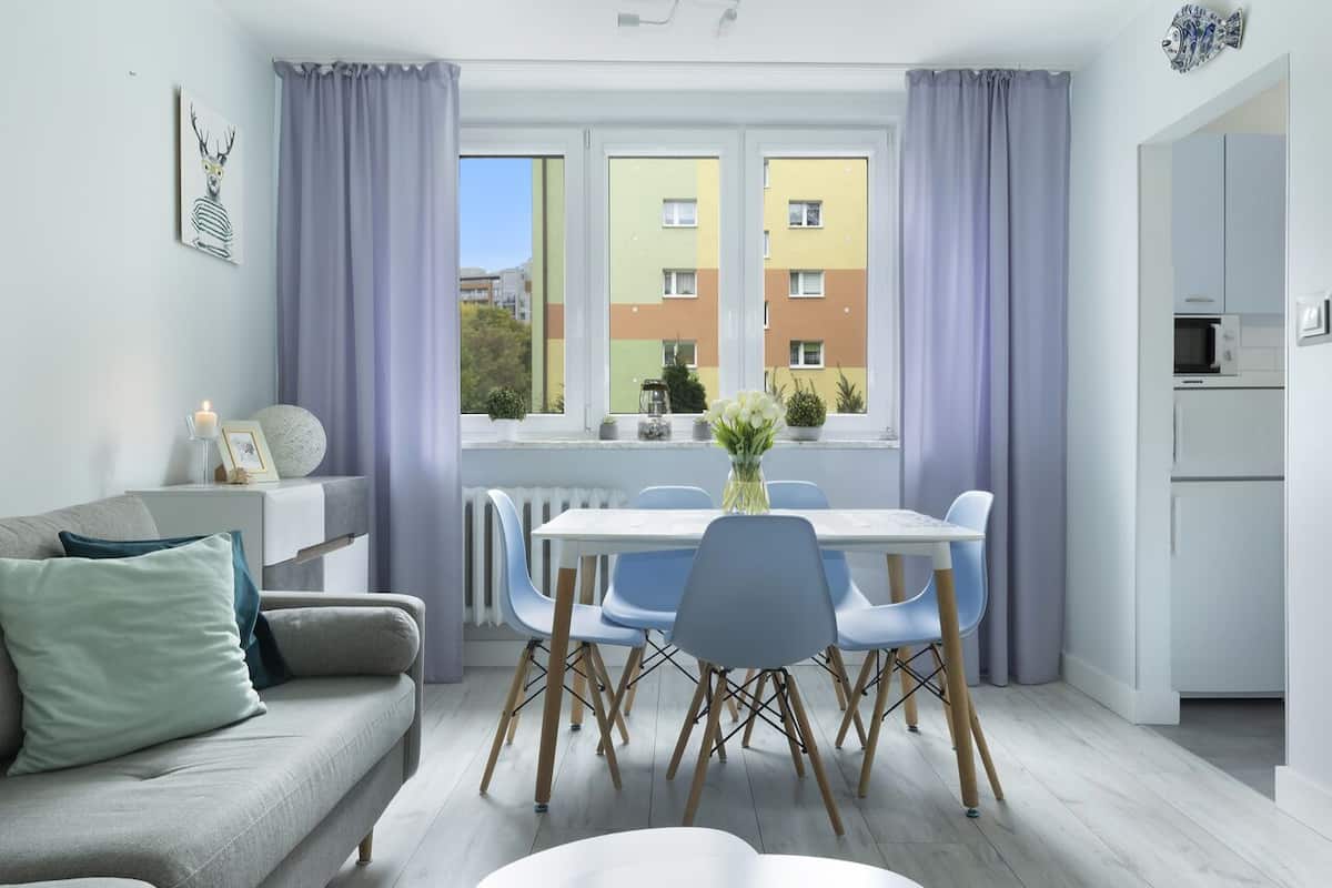A modern living room with a grey sofa, a dining table with blue chairs, and a view of colorful buildings through large windows.