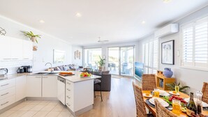 A modern kitchen with a central island, a dining area with wicker chairs, and a large window providing natural light. - Birubi First Wave, 45a Ocean Ave - pet friendly, waterfront, waterviews, air con, Wi-Fi (Anna Bay)