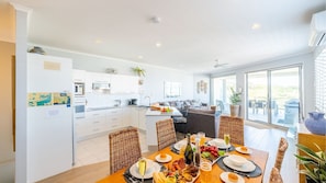 A modern kitchen with white cabinets, a dining area with a wooden table set for a meal, and a large sliding glass door leading to an outdoor patio. - Birubi First Wave, 45a Ocean Ave - pet friendly, waterfront, waterviews, air con, Wi-Fi (Anna Bay)