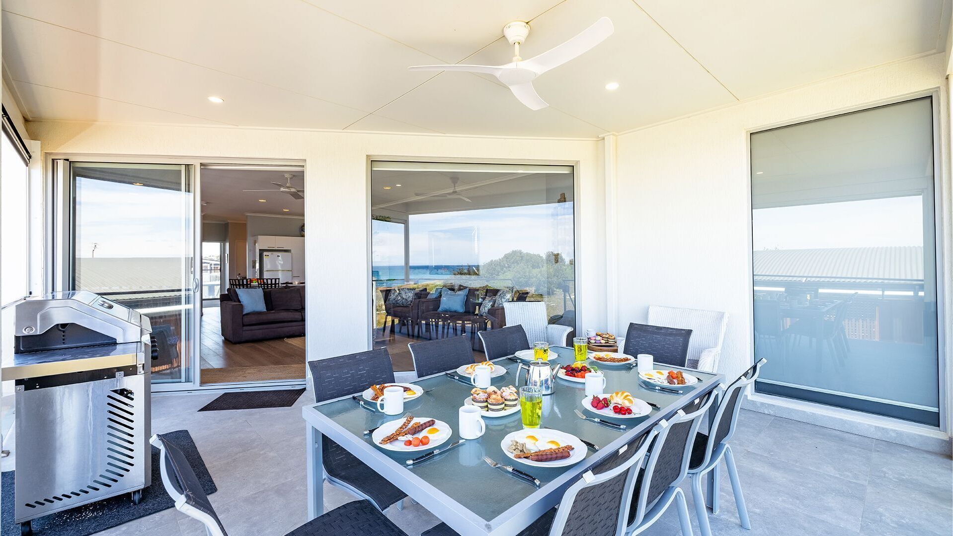 A modern dining area with a glass table set for breakfast, a grill, and a view of the sea.