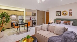 A compact hotel room with a kitchenette, a sofa, a dining table with chairs, and a bed with decorative pillows. - Prime Select Gamet El Dowal (Giza)