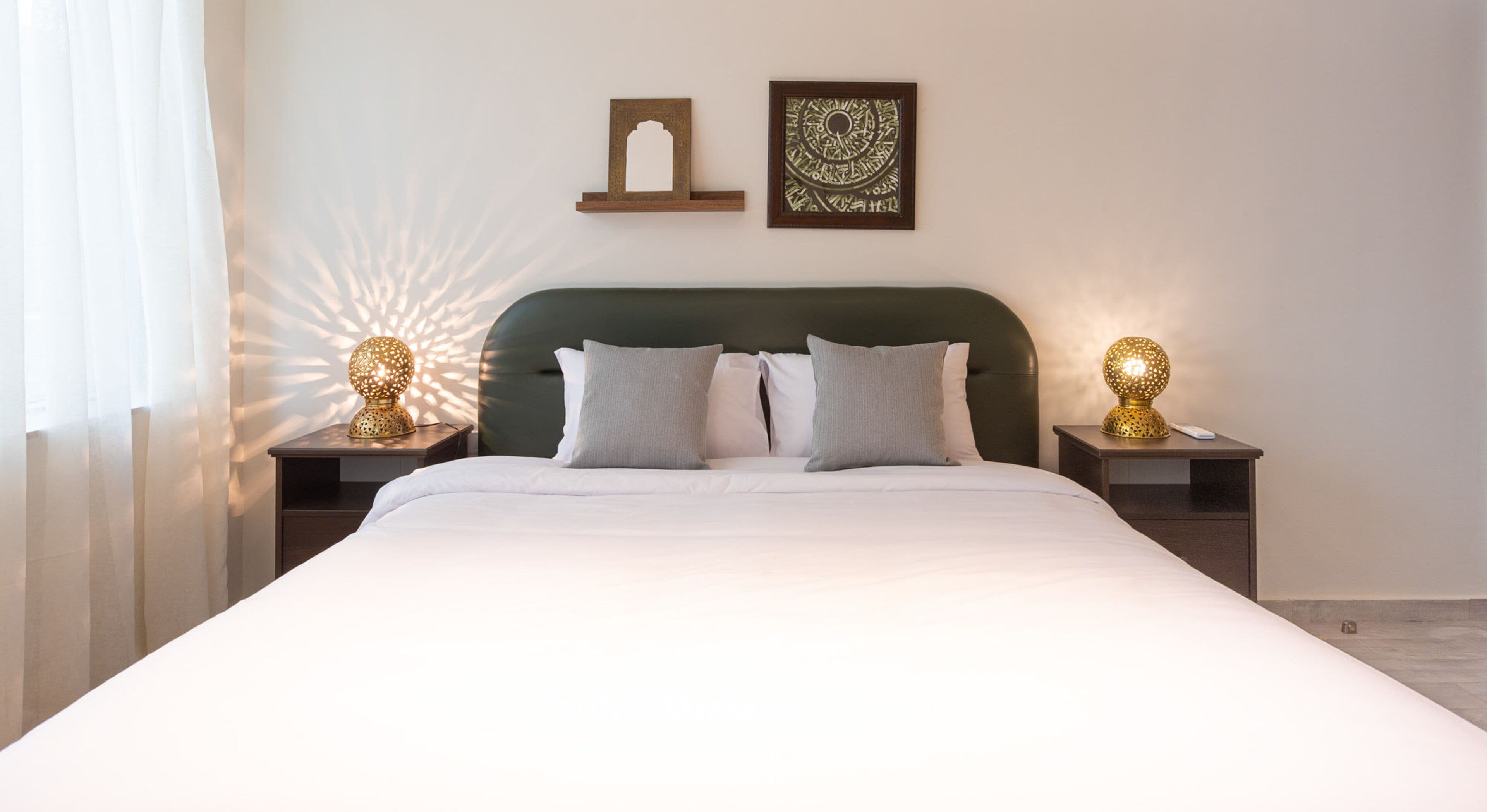 A neatly made bed with white linens, two bedside tables with decorative lamps, a wall-mounted shelf with a framed picture, and a window with sheer curtains allowing natural light.