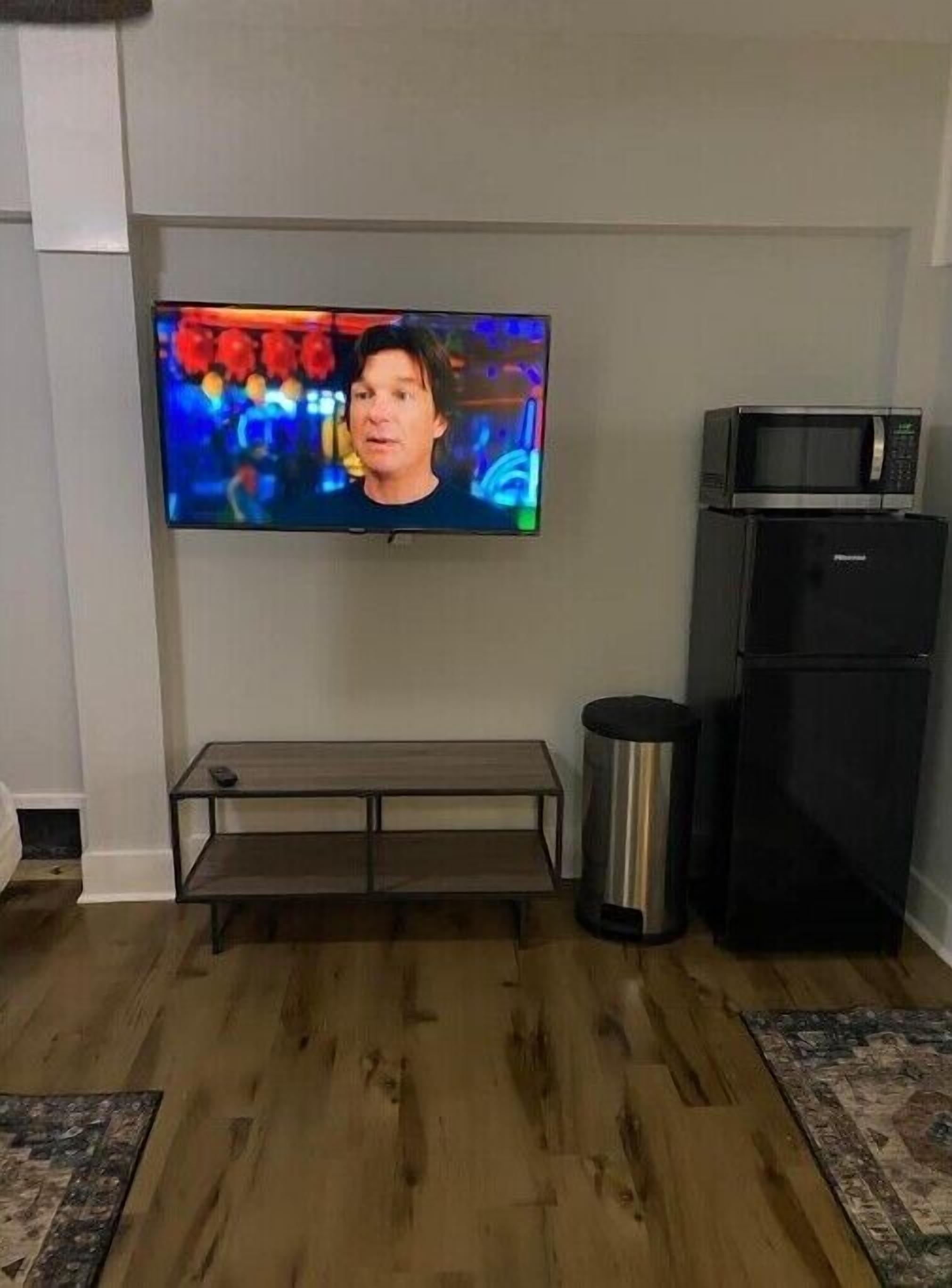 A living room with a flat-screen TV mounted on the wall, a black refrigerator, a metal trash can, and a wooden bench.