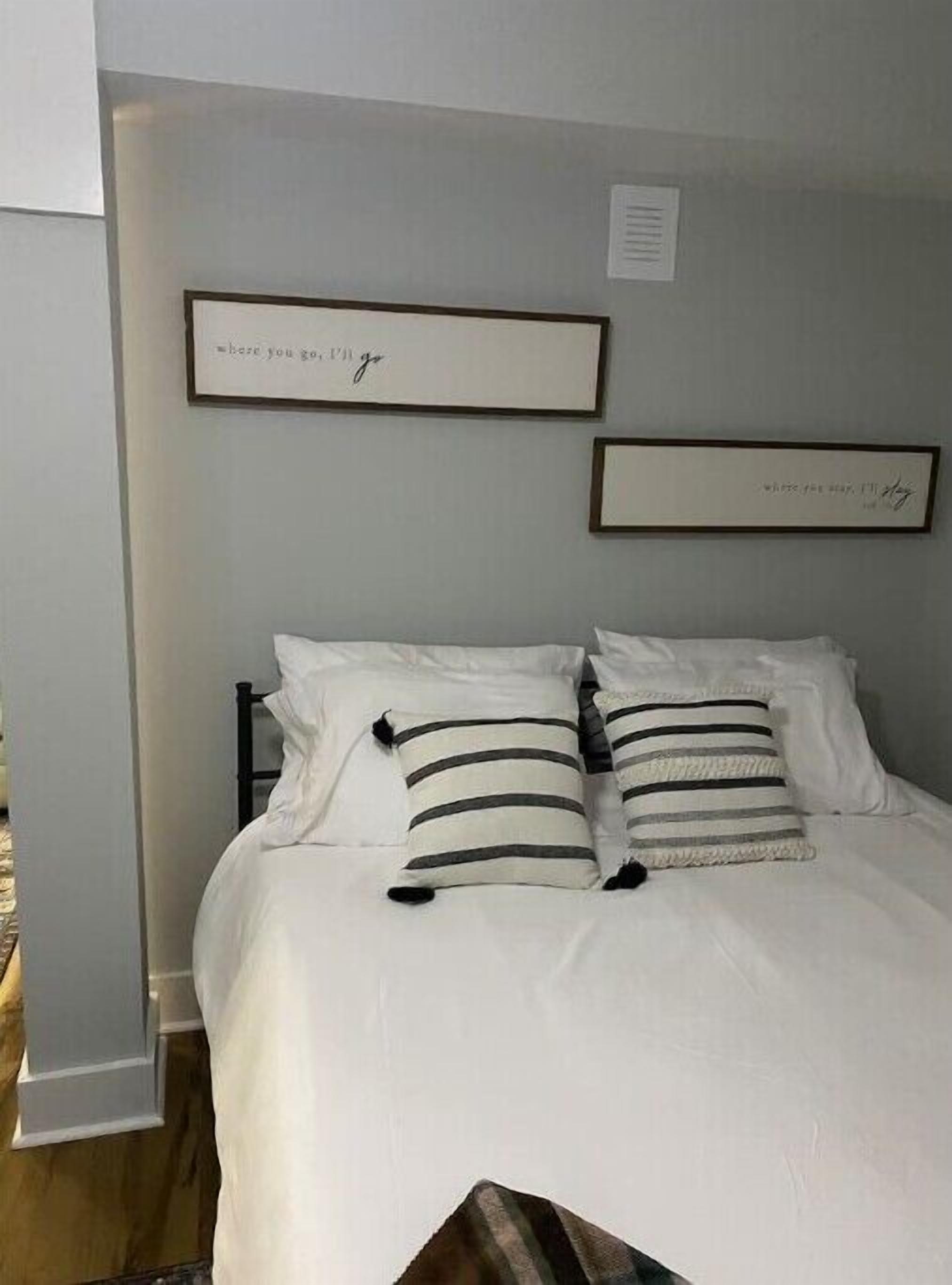 A neatly made bed with white bedding and striped pillows, a framed quote on the wall, and a small window with a view.
