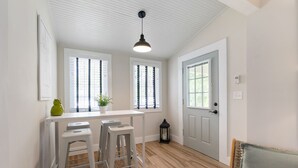 A bright, well-lit room with a white ceiling, large windows with blinds, a white door, and a small table with stools.