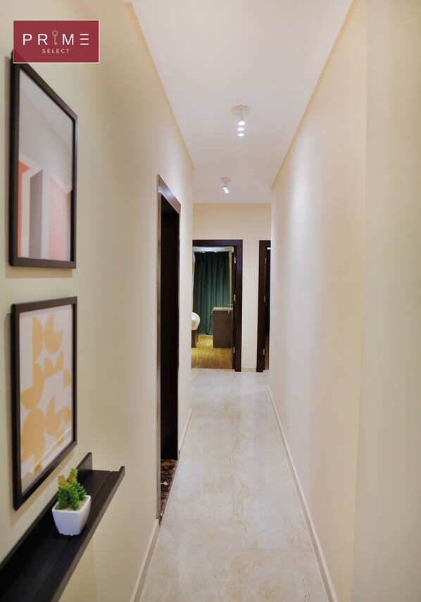 A hallway with framed artwork on the walls, a potted plant, and a view into a room with a bed and curtains.