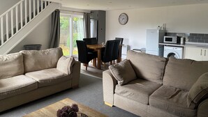 A living room with two sofas, a dining table with chairs, a staircase, a kitchen with appliances, and a clock on the wall. - Cosy Cotswold Coachhouse - Rural and quiet (Chalford)