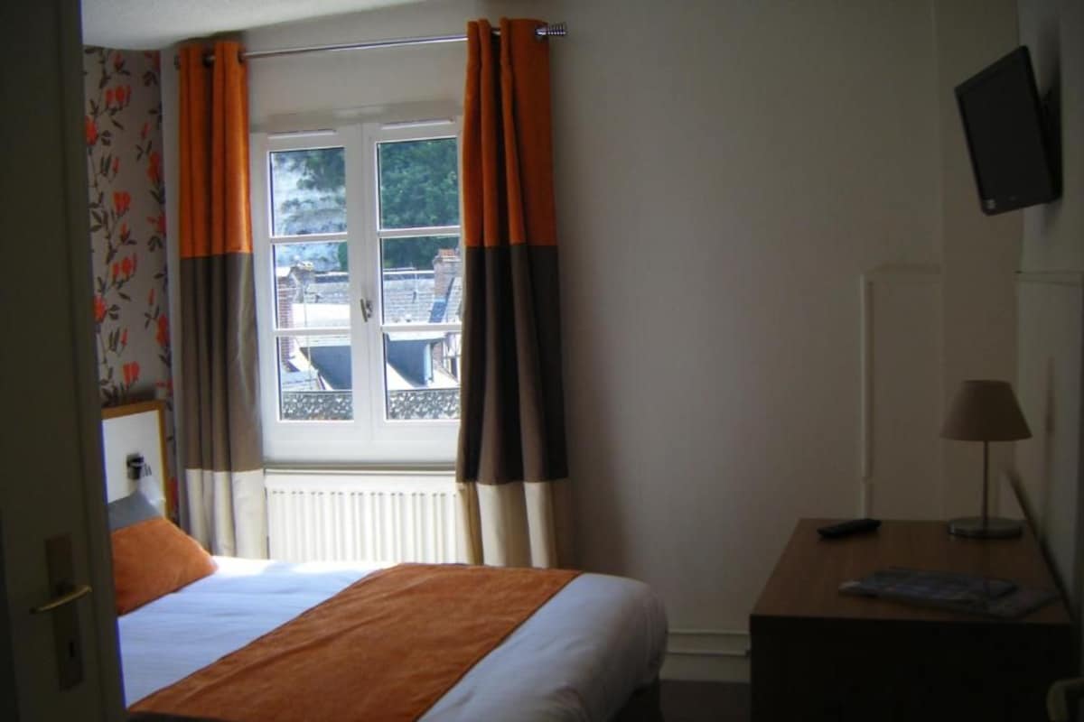 Quarto de hotel com cama, escrivaninha, televisão e janela com cortinas.