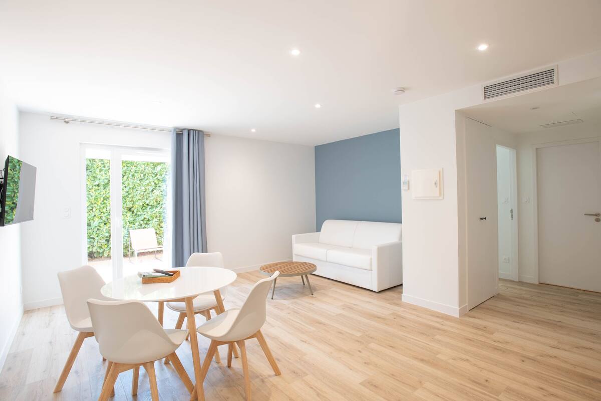 A modern living room with a white sofa, a dining table with chairs, and a large window with a view of greenery.