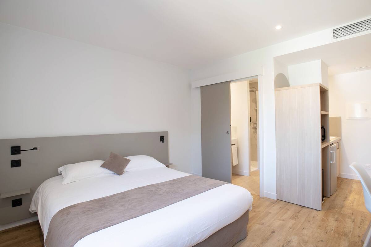 A modern hotel room with a large bed, a bedside table, a wardrobe, and a bathroom visible through an open door.