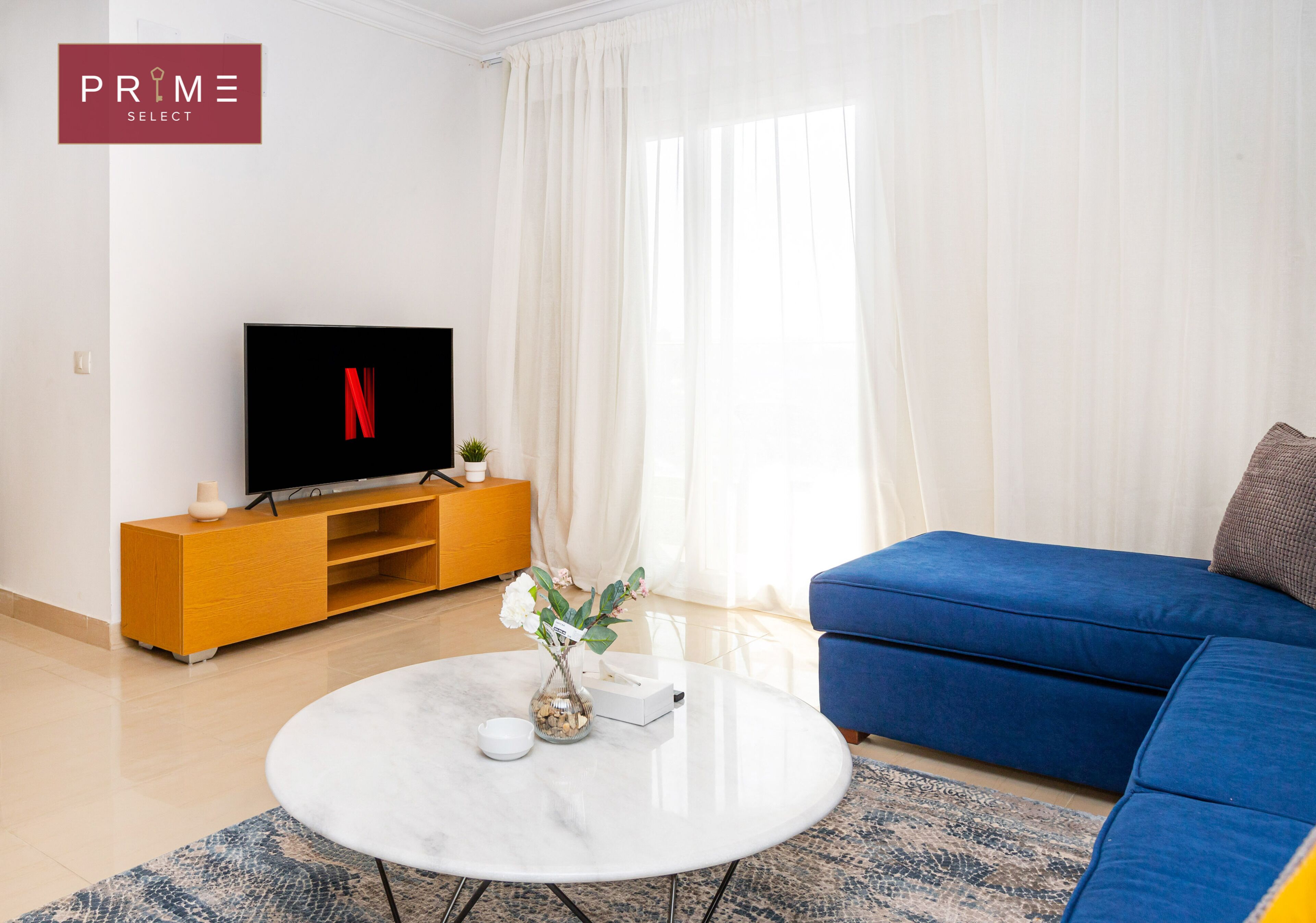 Ein modernes Wohnzimmer mit einem Flachbildfernseher auf einem Holz-TV-Möbel, einem blauen Sofa, einem weißen Couchtisch und einem großen Fenster mit durchsichtigen Vorhängen.
