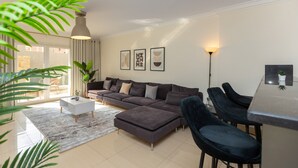 Ein modernes Wohnzimmer mit grauem Sofa, einem weißen Couchtisch und einem dunklen Hocker. Es gibt einen Glastisch mit zwei Stühlen, eine Zimmerpflanze und eine Stehlampe.