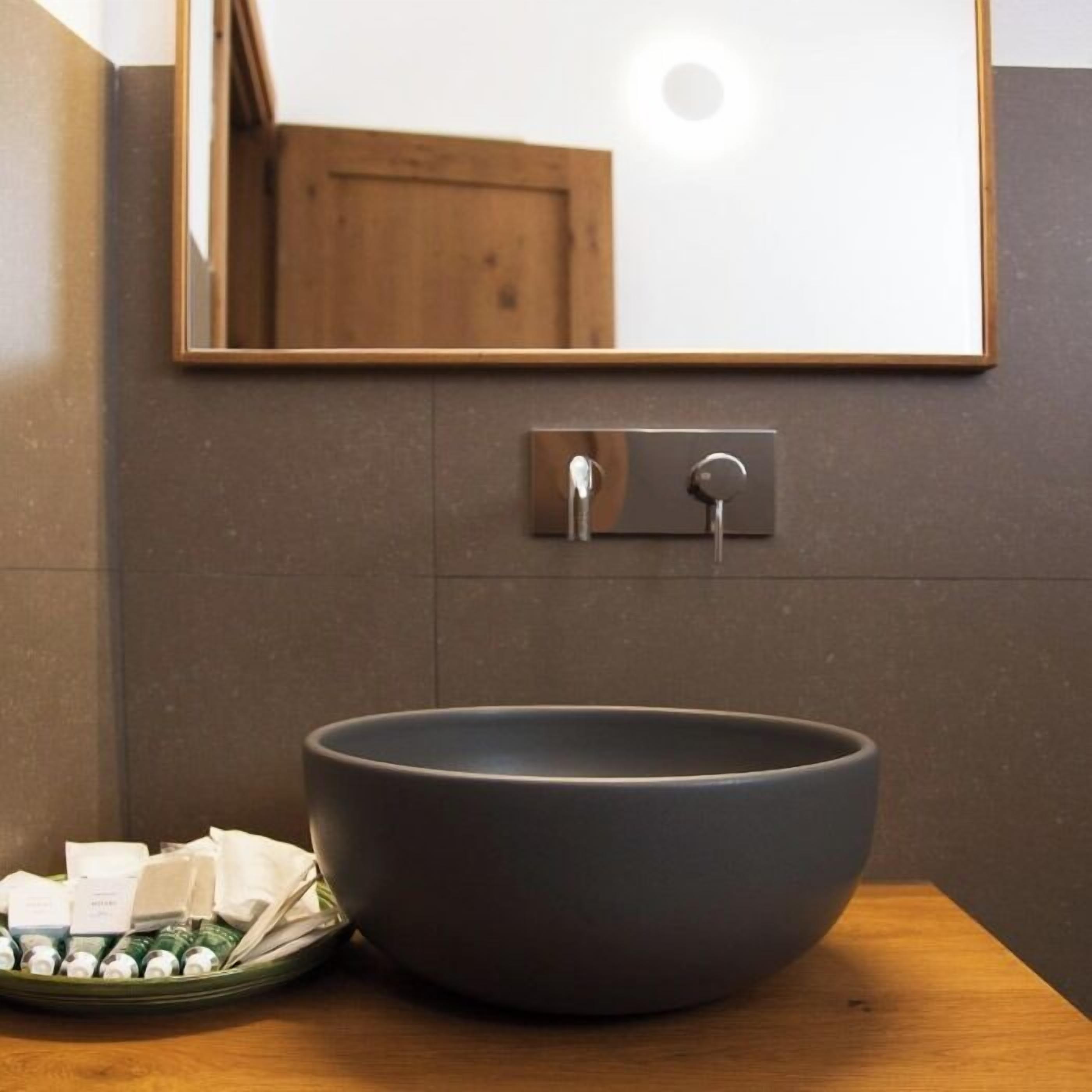 Une salle de bain dotée d’un grand lavabo de couleur sombre, d’un plan de toilette en bois et d’un miroir placé au-dessus du lavabo.