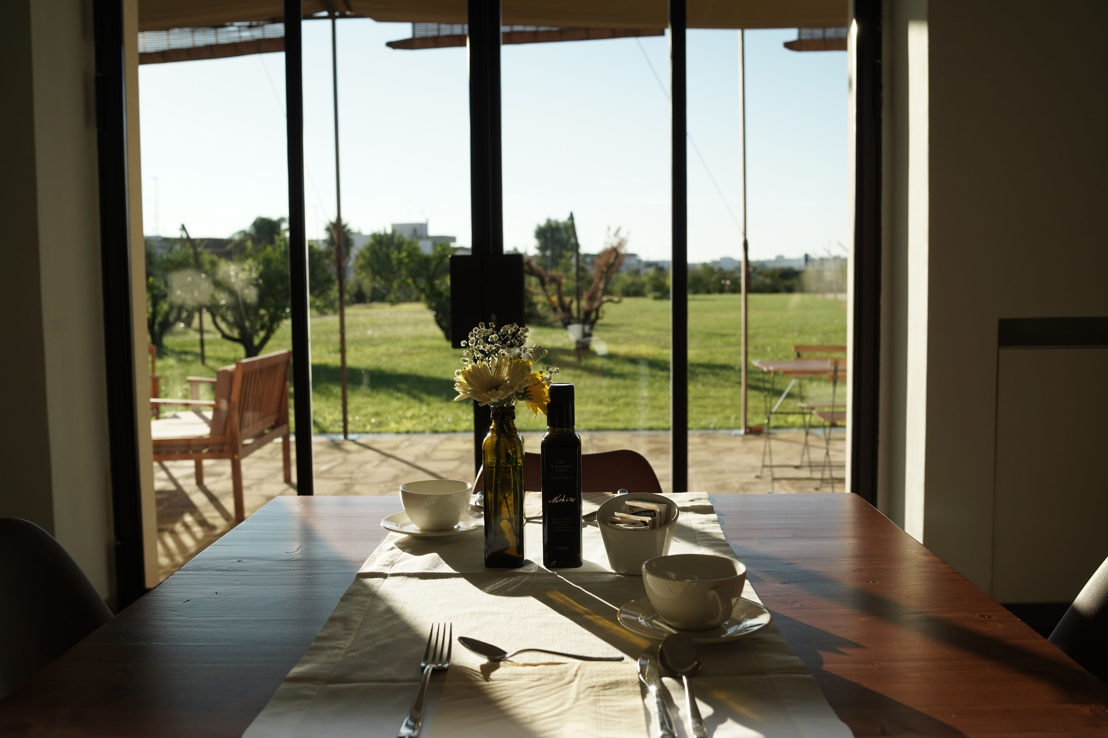A dining table set with a vase of flowers, a bottle, and a cup, with a view of a grassy outdoor area through large windows.