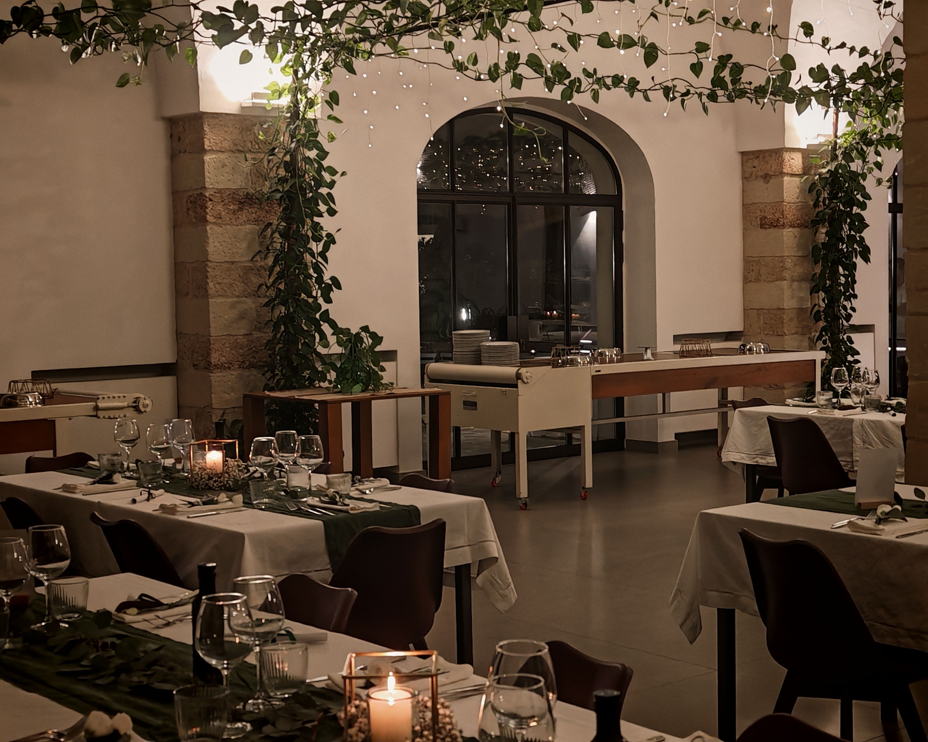 A restaurant with tables set for dining, a large arched window, and hanging plants.