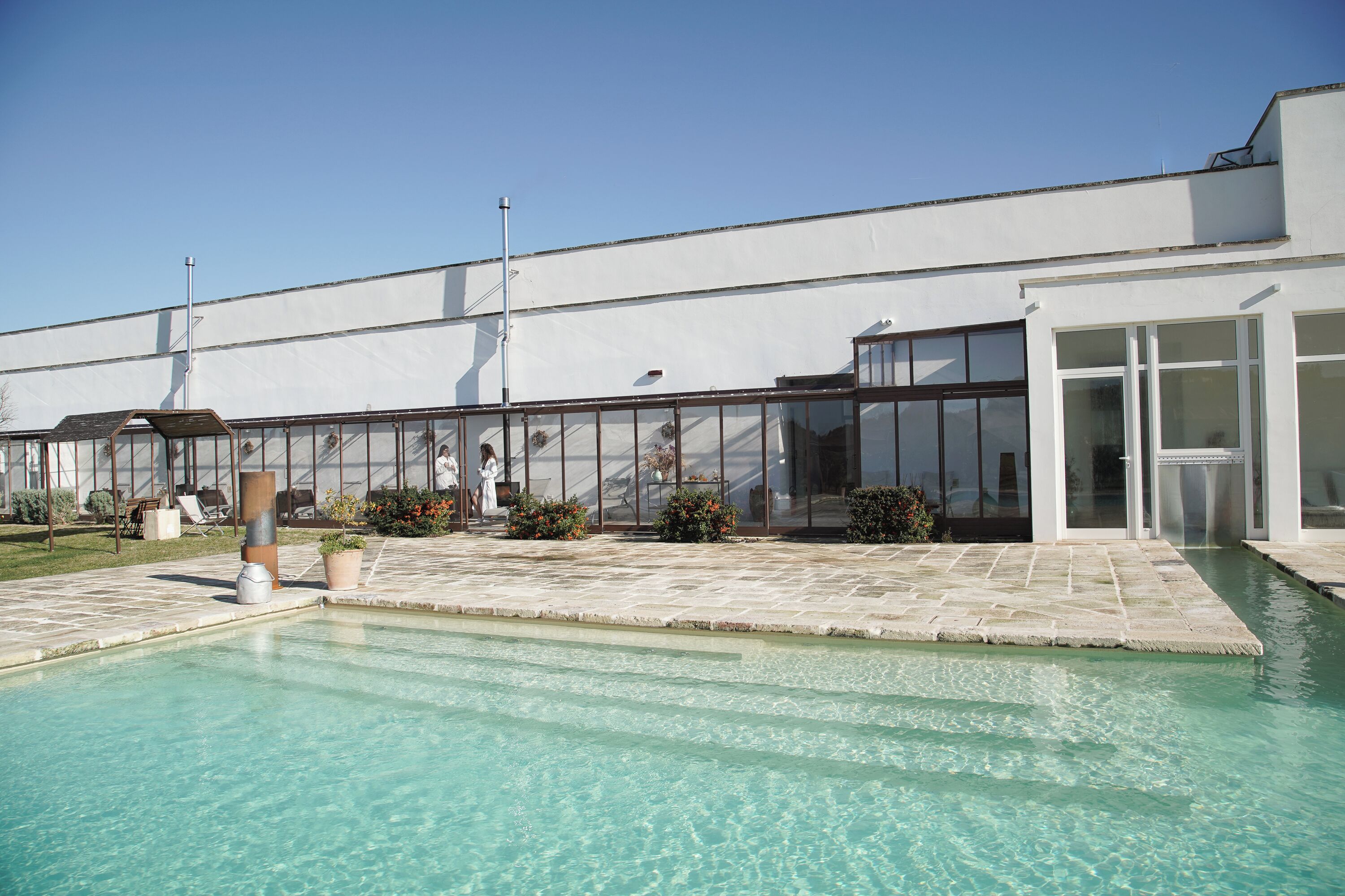 A swimming pool area with a clear pool, a white building with large windows, and outdoor seating.
