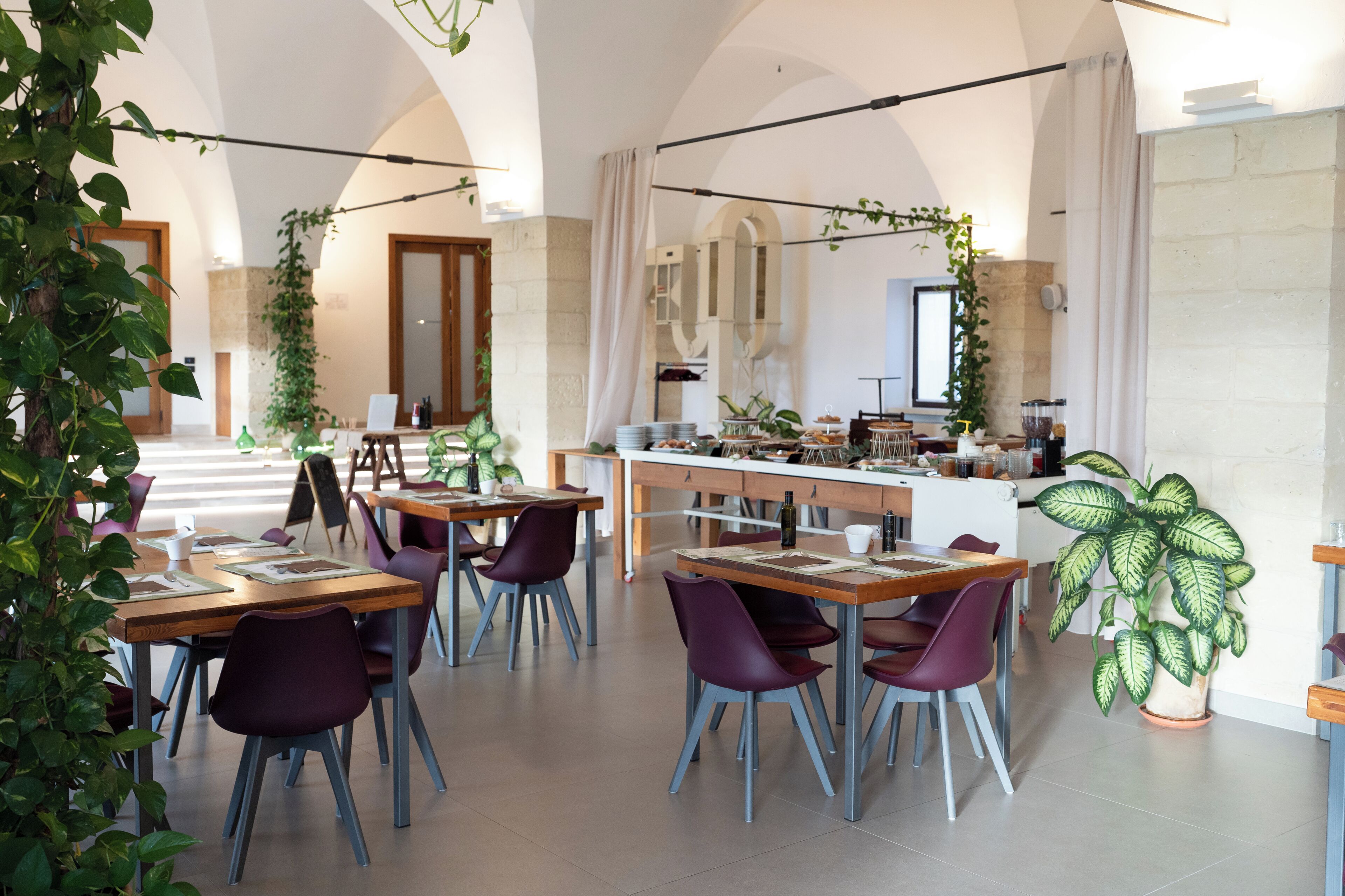 A spacious dining area with tables and chairs, a kitchen in the background, and potted plants.
