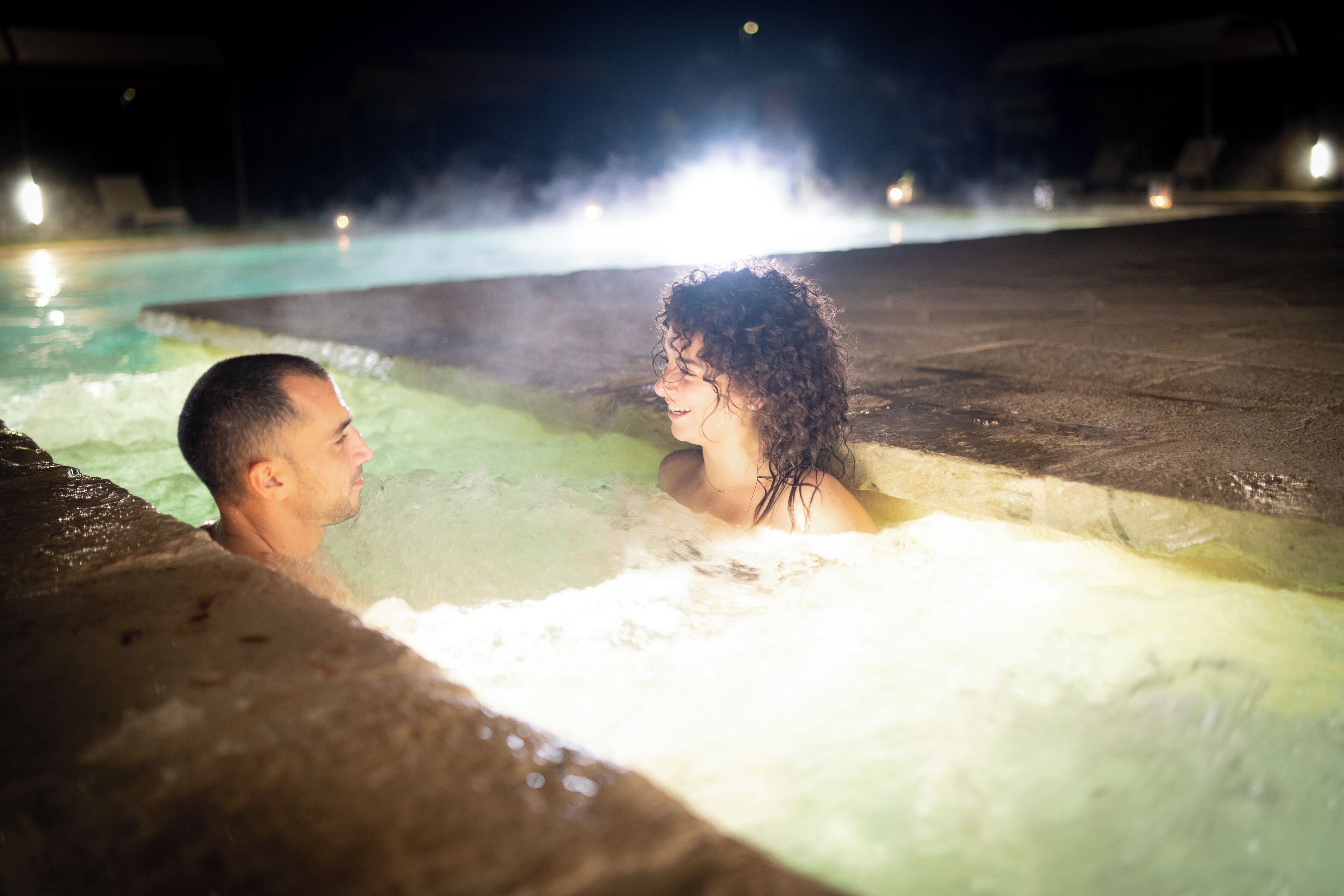 A couple in a hot tub at night with a pool in the background.