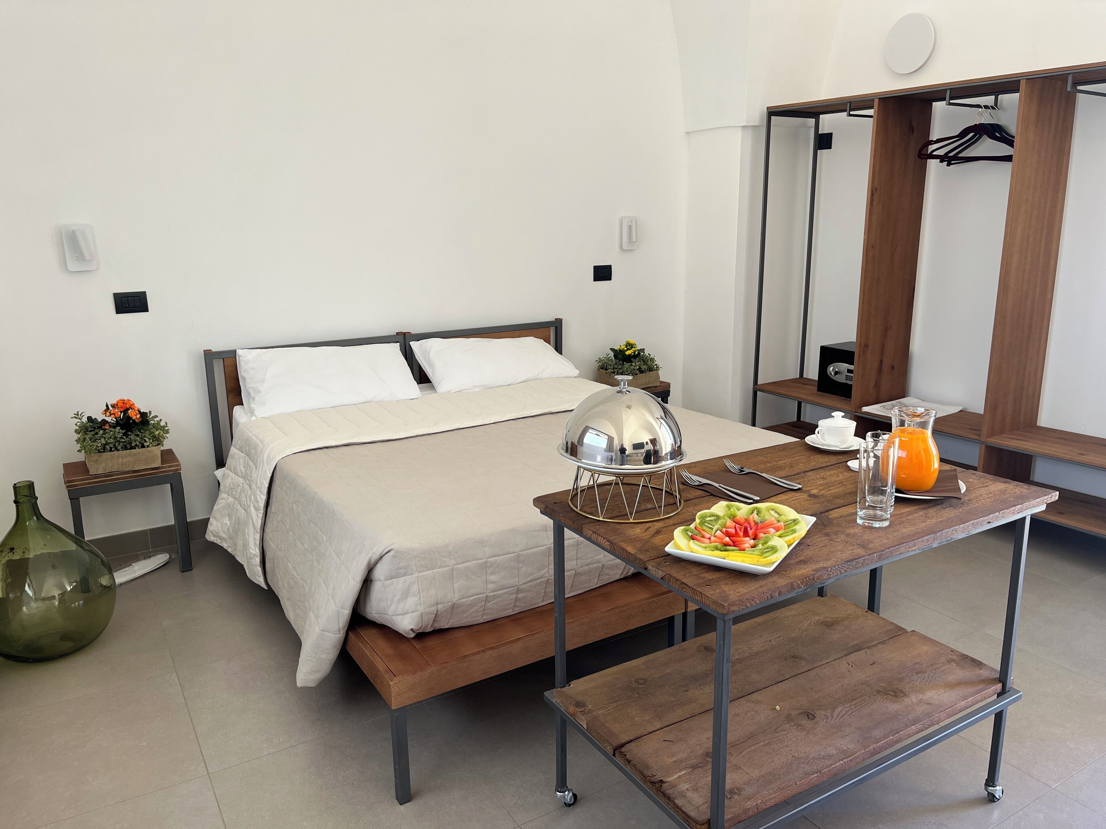 Habitación de hotel impecable con una cama, mesitas de noche, una mesa de desayuno con una bandeja de fruta, una jarra de jugo y un espejo.