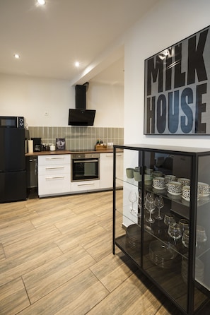 Une cuisine moderne avec un réfrigérateur noir, des armoires blanches, une vitrine contenant des assiettes et des verres à vin, et une enseigne « LAITERIE » accrochée au mur.
