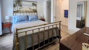Una camera da letto con un letto, una ventola a soffitto, un quadro raffigurante una spiaggia, un pavimento in legno e un bagno visibile attraverso una porta aperta.