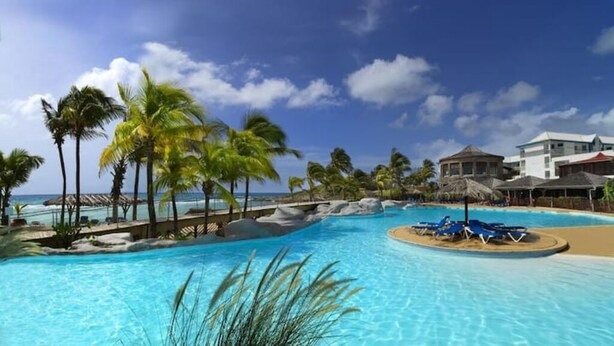 Une piscine d’un complexe hôtelier avec une eau d’un bleu limpide, des palmiers et un espace aménagé en plage.