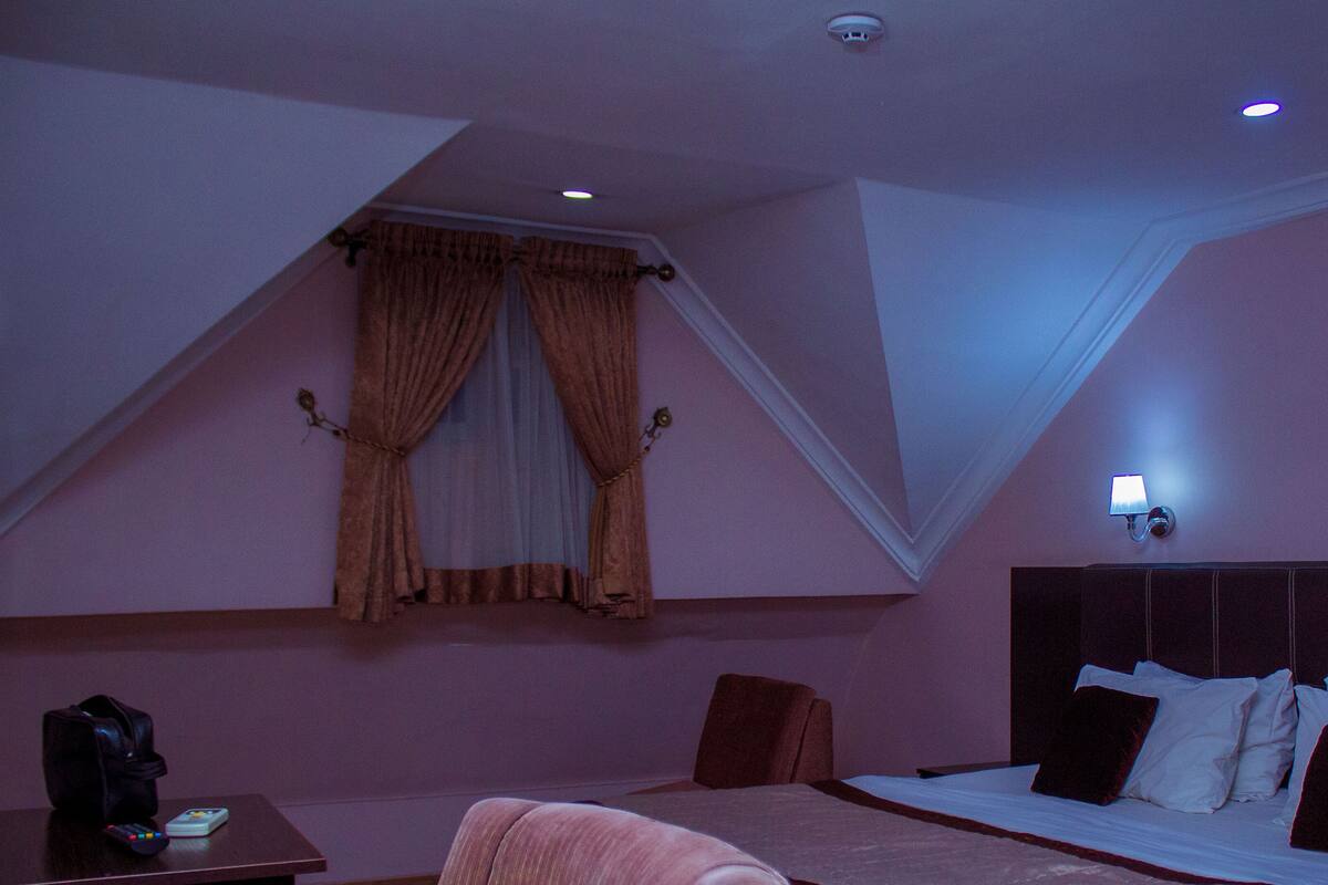 A bedroom with a sloped ceiling, a bed with white bedding, a nightstand with a remote control, and a wall-mounted lamp.