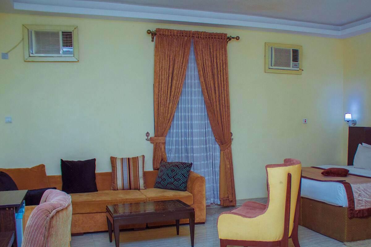 A room with a sofa, a chair, a bed, and a table. There are two air conditioning units on the walls.