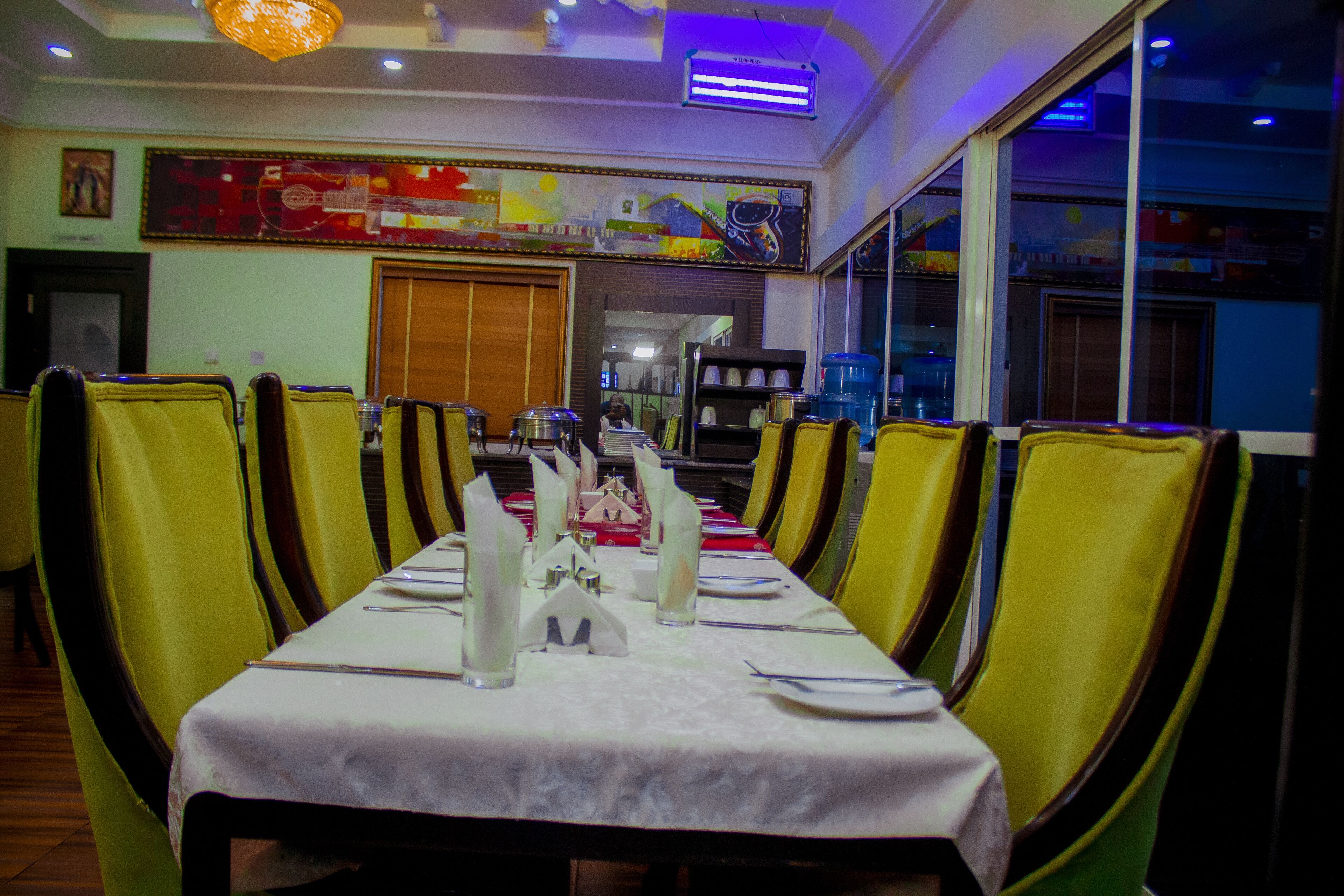 A restaurant with a long table set for dining, yellow chairs, and a decorative ceiling light.