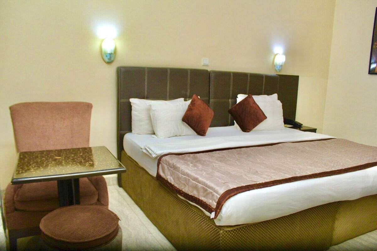 A hotel room with a bed, a chair, a small table, and two wall-mounted lamps.