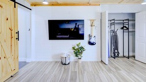 A room with a wooden floor, a television mounted on the wall, a guitar, a plant, and a metal rack with clothes. - Modern Studio with King Bed, Free Parking & Wi-Fi (Memphis)