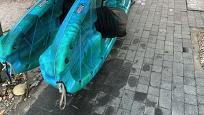 Un kayak azul con reposapiés negros y una bolsa negra sujeta, estacionado bajo una zona cubierta con un árbol y una casa al fondo.