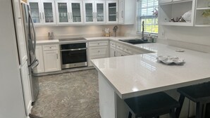 Cocina moderna con muebles blancos, horno de acero inoxidable, encimera de mármol y ventana con vistas.