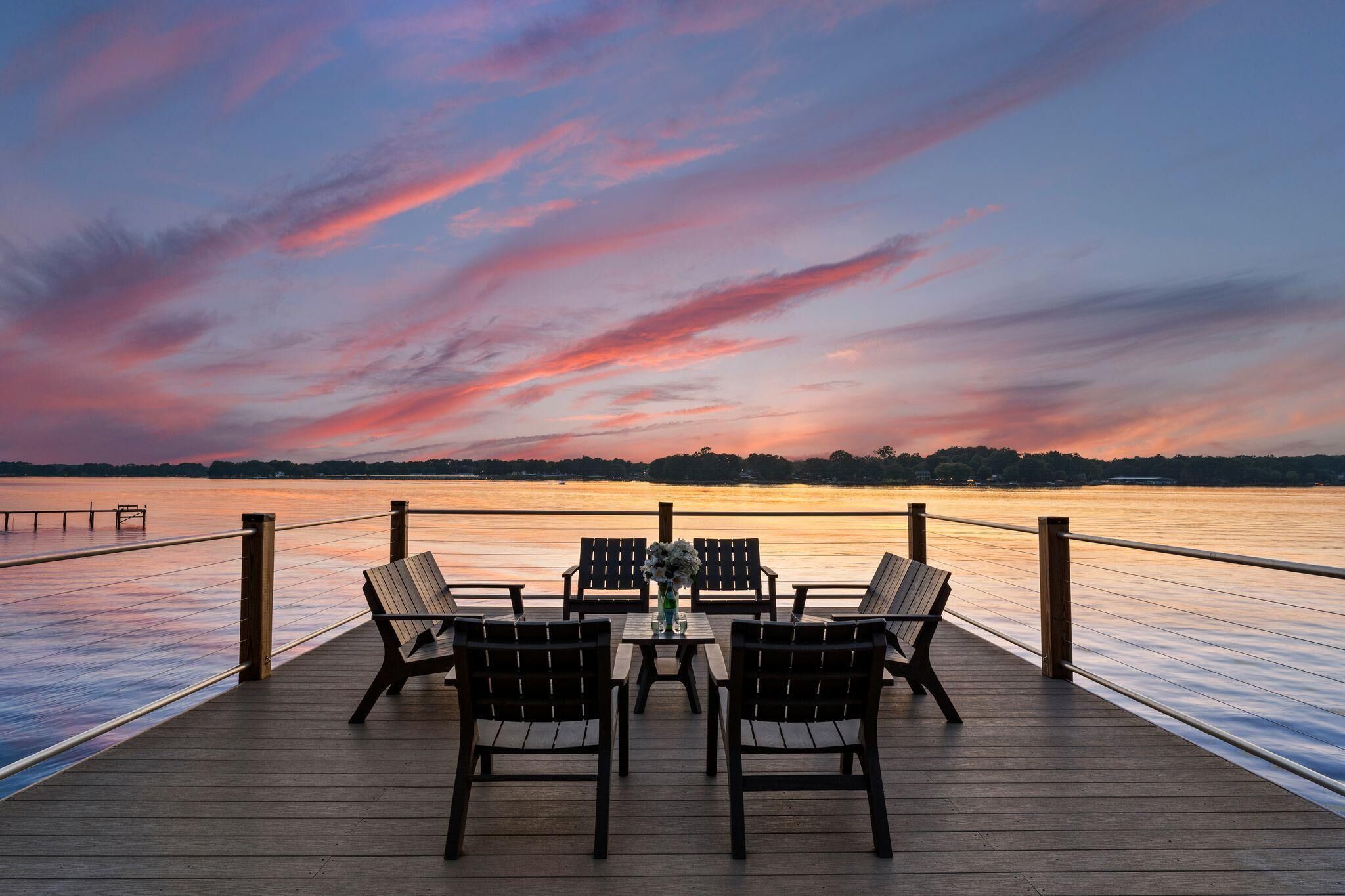 A wooden deck with a table and chairs facing a calm body of water during sunset.
