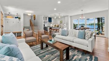 A spacious living room with a large sofa, two armchairs, and a coffee table. A kitchen is visible in the background, and there is a balcony with a view of the lake.