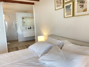 Un dormitorio con cama, mesita de noche, lámpara de pared y baño visible a través de una puerta abierta.